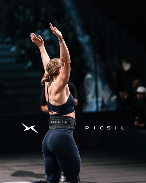 An athlete stands with arms raised in victory, showcasing the weightlifting belt as a badge of hard-won achievement in a dark, dramatic arena setting.
